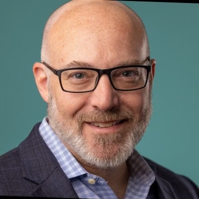 Smiling bald man with glasses and a beard in a navy blazer, teal background